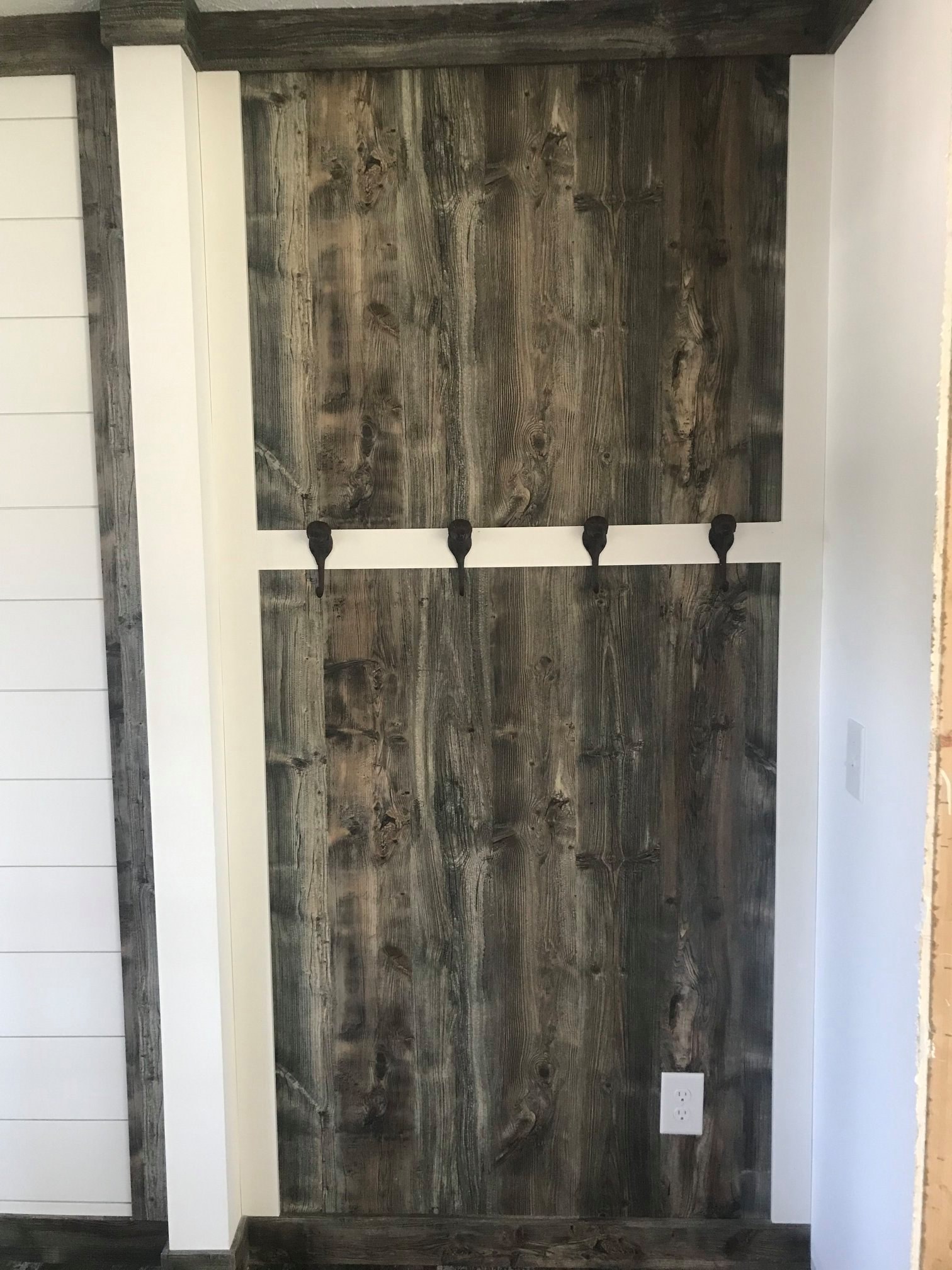 A rustic wooden wall features four dark metal hooks, set in a cozy alcove with a white frame. The wood’s texture adds warmth and character.