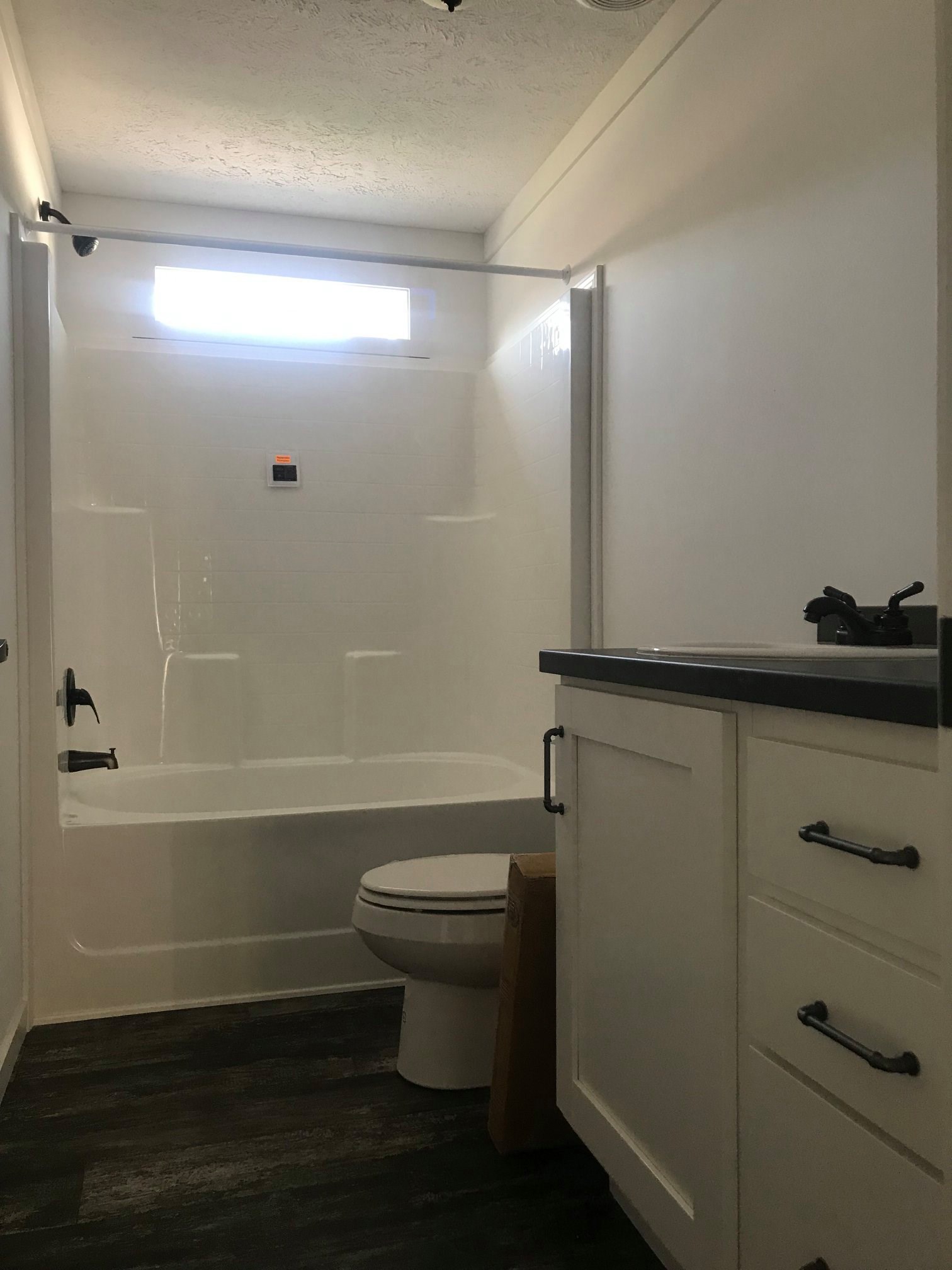 A modern bathroom features a white bathtub with an overhead shower, a small window above, a toilet, and a white vanity with a dark countertop, creating a clean and minimalist atmosphere.