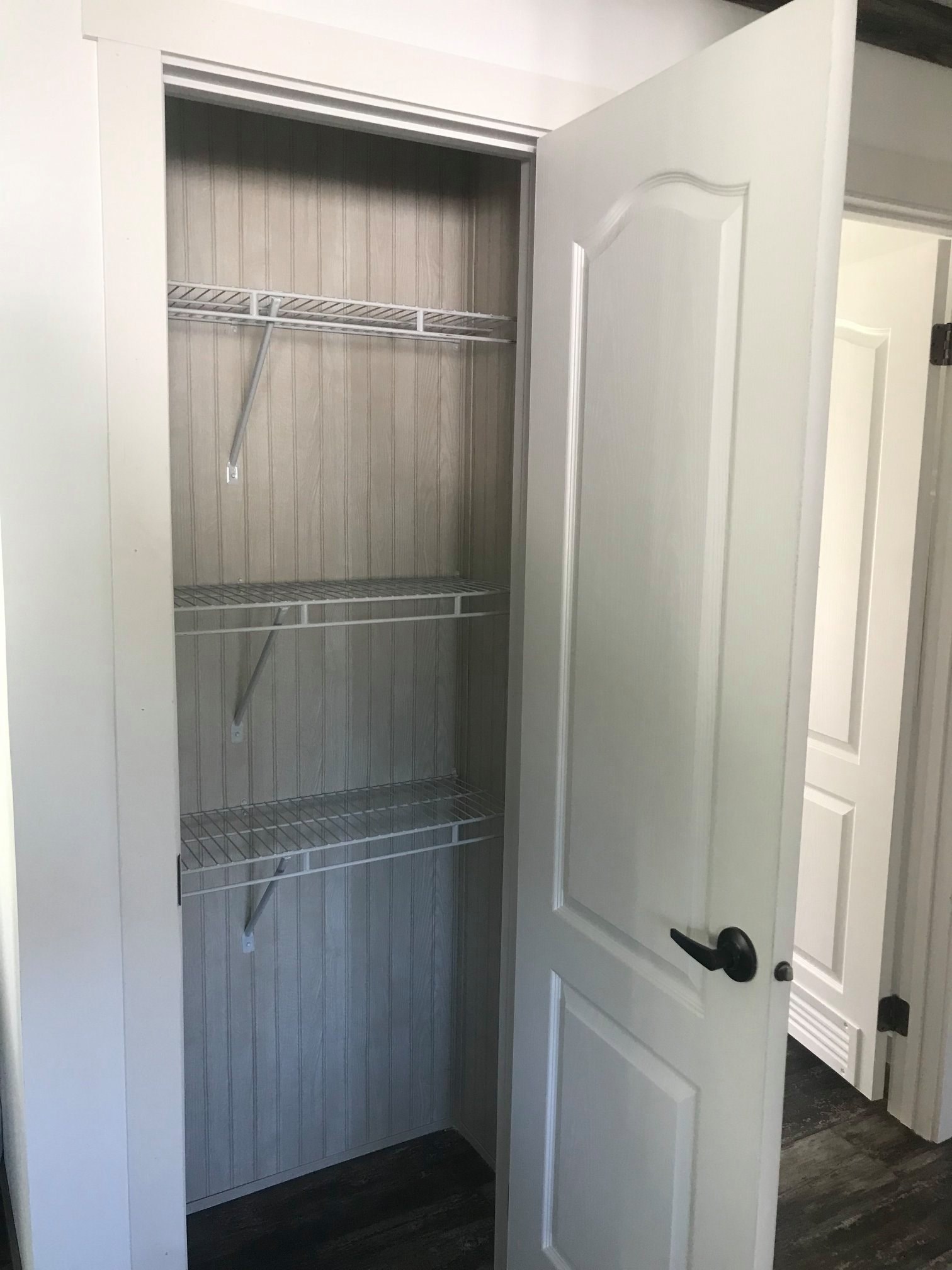 Open white closet with wire shelves and wood panel backing. Door partially open, revealing dark wooden floor. Neutral, minimalist tone.