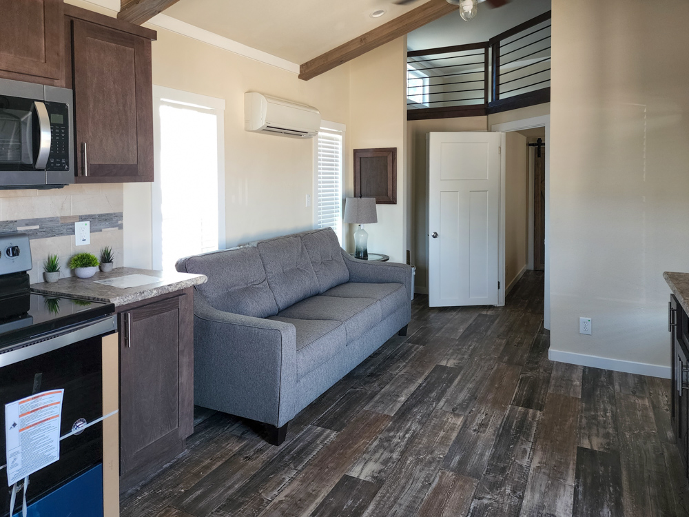 A modern tiny home interior featuring a gray couch, wooden cabinets, and a compact kitchen with a microwave. The space is bright, with wooden floors.