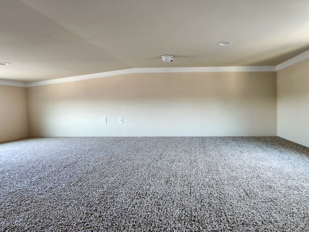 Low-ceilinged loft space with beige walls and plush carpet. Two outlets are visible on the back wall, and a smoke detector is mounted on the ceiling.