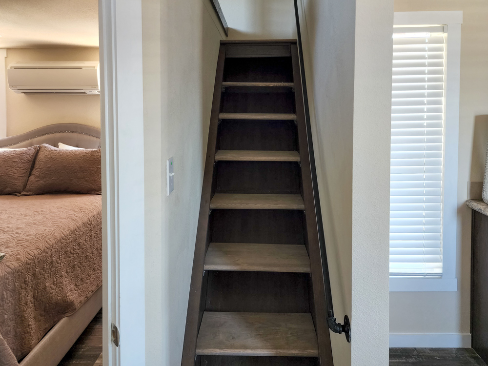 Narrow wooden staircase leading up between two walls. On the left, a bedroom with a bed and air conditioner; on the right, a window with closed blinds.