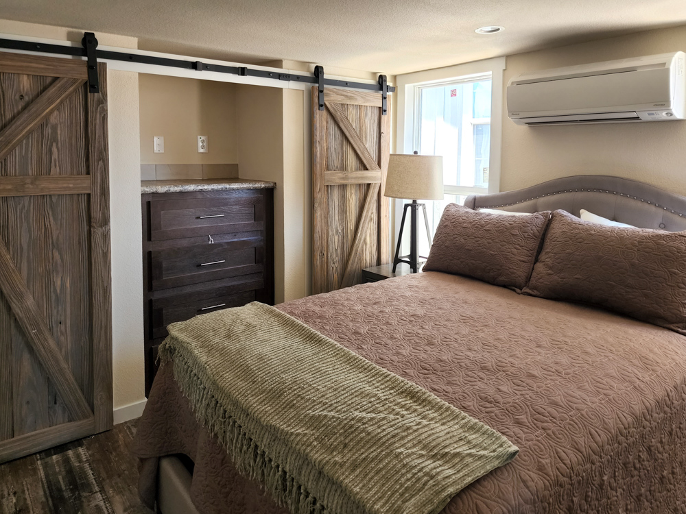 Cozy bedroom with a brown quilted bedspread, wooden barn-style closet doors, a dark wooden dresser, green throw, and soft lighting.