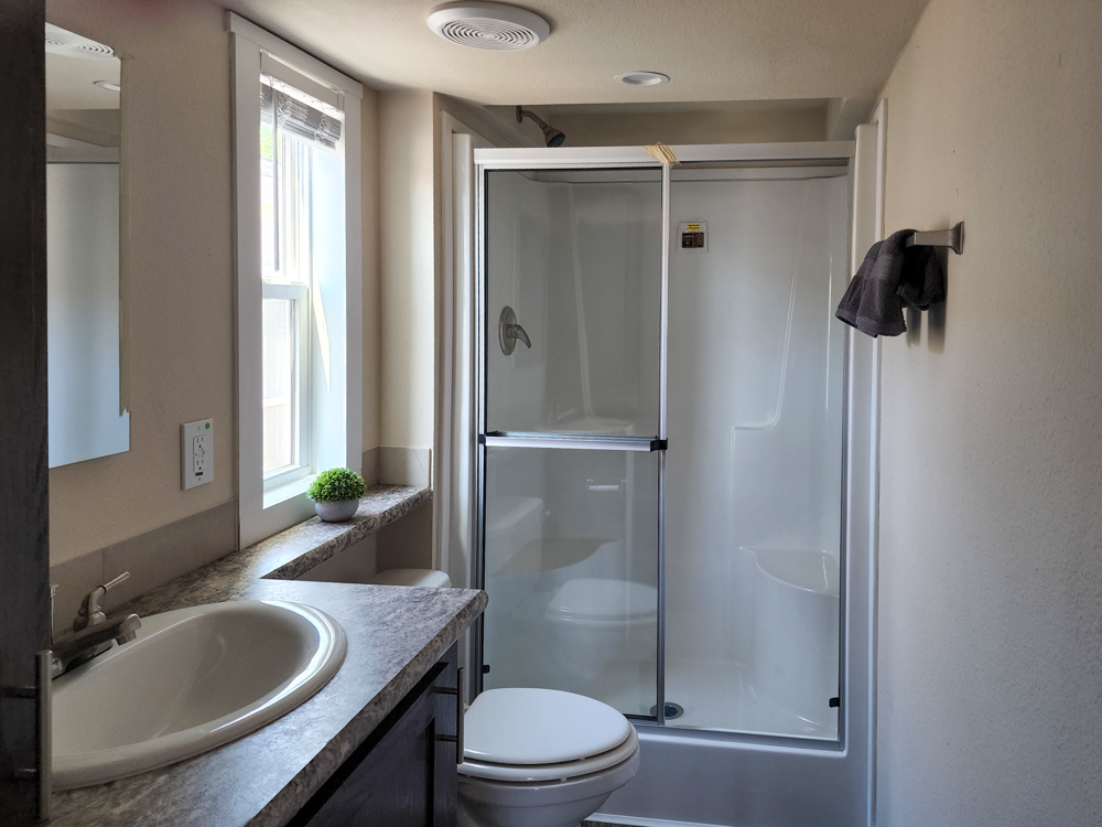 A small, well-lit bathroom with a white sink, marble countertop, and glass shower. A towel hangs on the wall, and a small plant sits by the window.