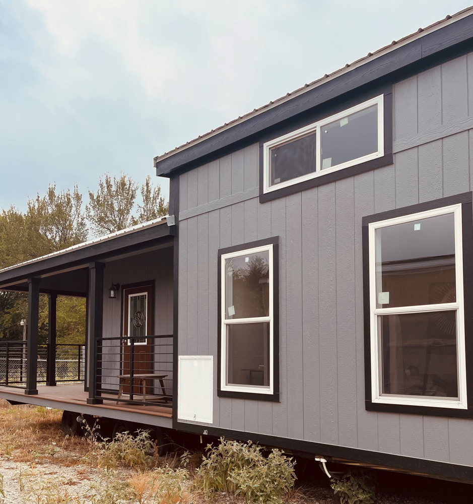 Modern tiny house with gray siding and white-trimmed windows. A wooden porch is in front, set against a backdrop of trees under a cloudy sky.
