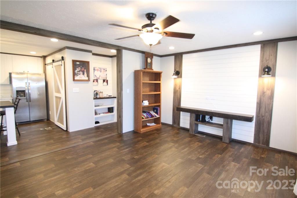 A modern living space with dark wood floors, a ceiling fan, and white walls. A built-in bookshelf and a shiplap accent wall create a cozy feel. The room is well-lit, with open access to a kitchen with stainless steel appliances and a sliding barn door.