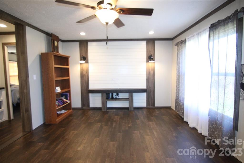 Cozy room with dark wood flooring, ceiling fan, and white shiplap accent wall with sconces. Wooden bookshelf and sheer curtains add warmth.