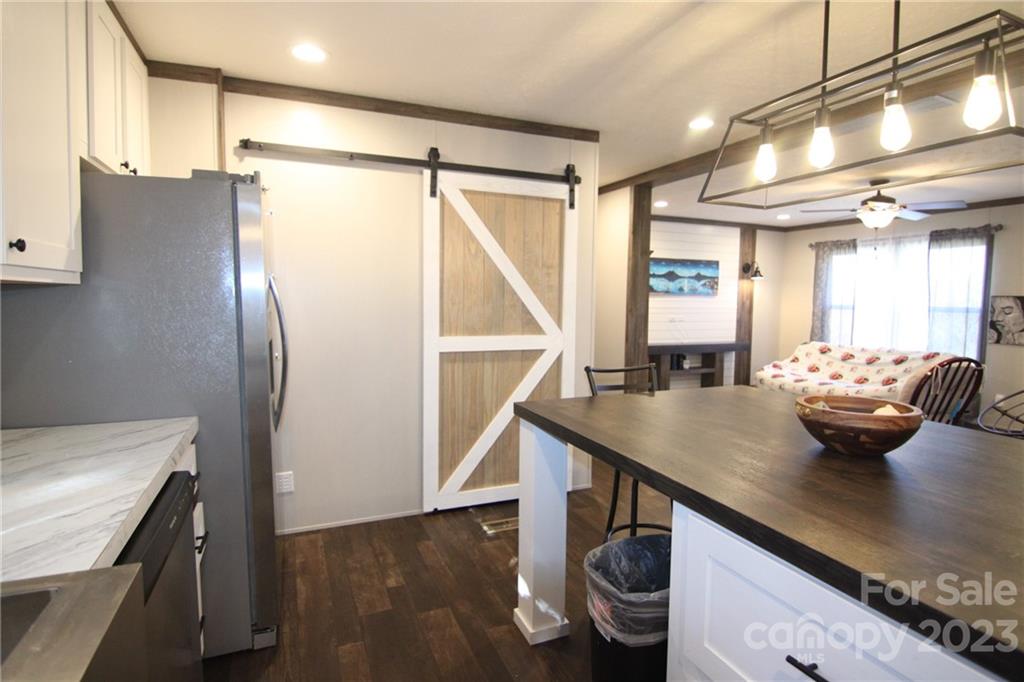 Modern kitchen with dark wood floors, a white island, and stainless steel appliances. A rustic sliding barn door leads to a cozy living area with a fan.
