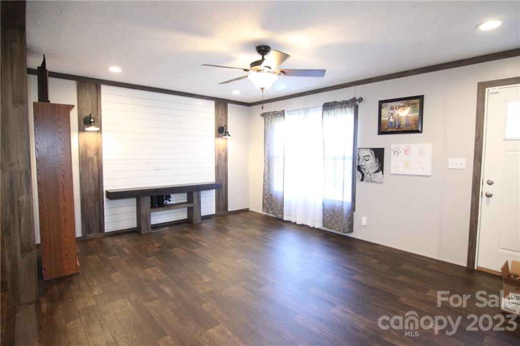A spacious living room with dark wooden floors, light walls, and a ceiling fan. A large window with sheer curtains allows natural light. Minimalist decor includes wall lamps and artwork.