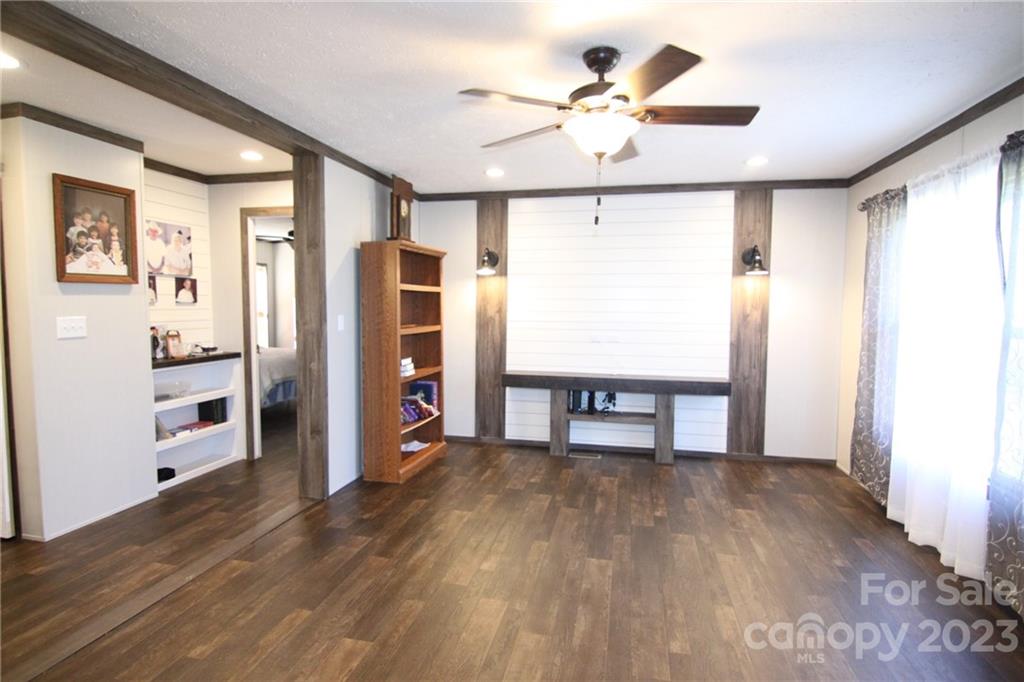 A cozy living room with dark wood flooring, a ceiling fan, and a shiplap accent wall. Shelves on the left display photos and decorations.