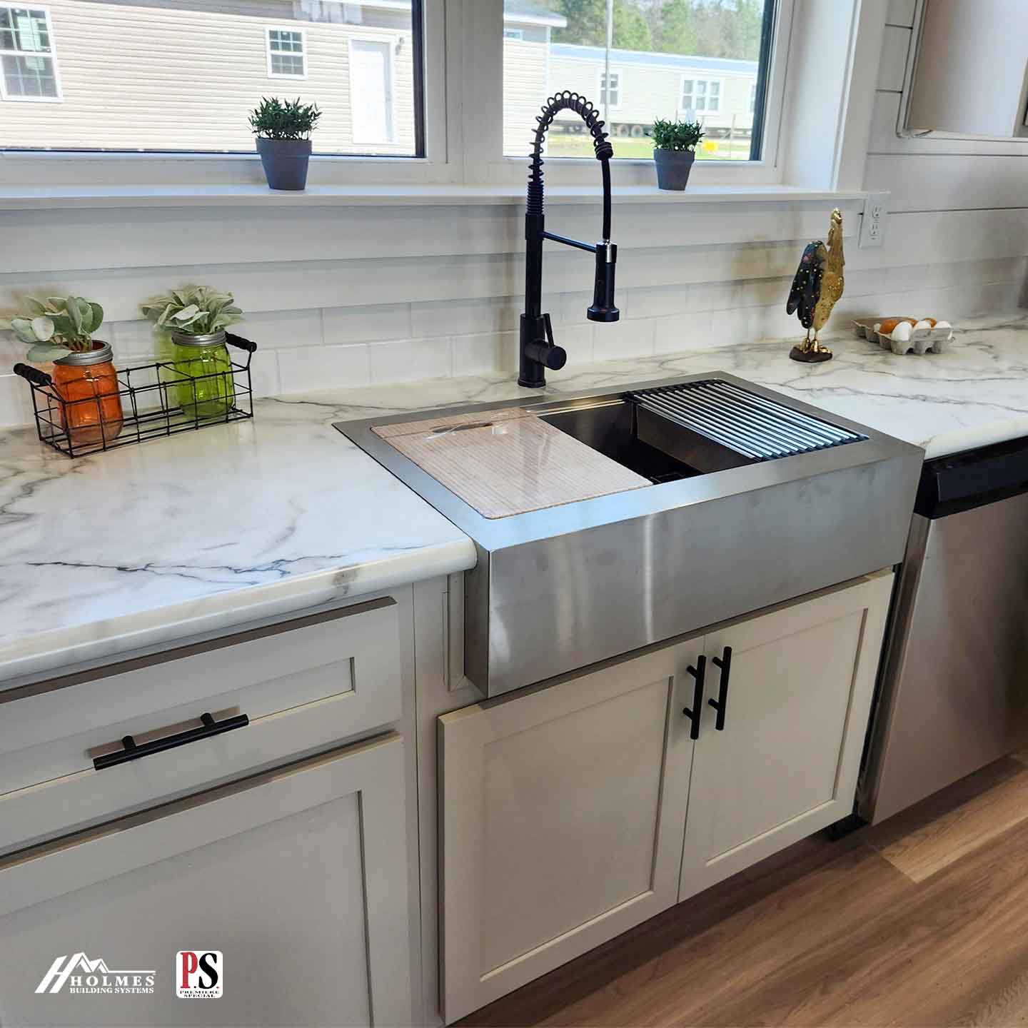 Modern kitchen with a stainless steel farmhouse sink, white marble countertop, and black faucet. Nearby are jars with plants, a rooster figurine, and eggs.