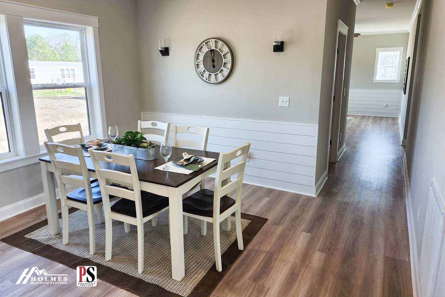 A cozy dining area with a wooden table and six chairs on a woven rug. Neutral walls, a round clock, and soft natural light create a welcoming atmosphere.