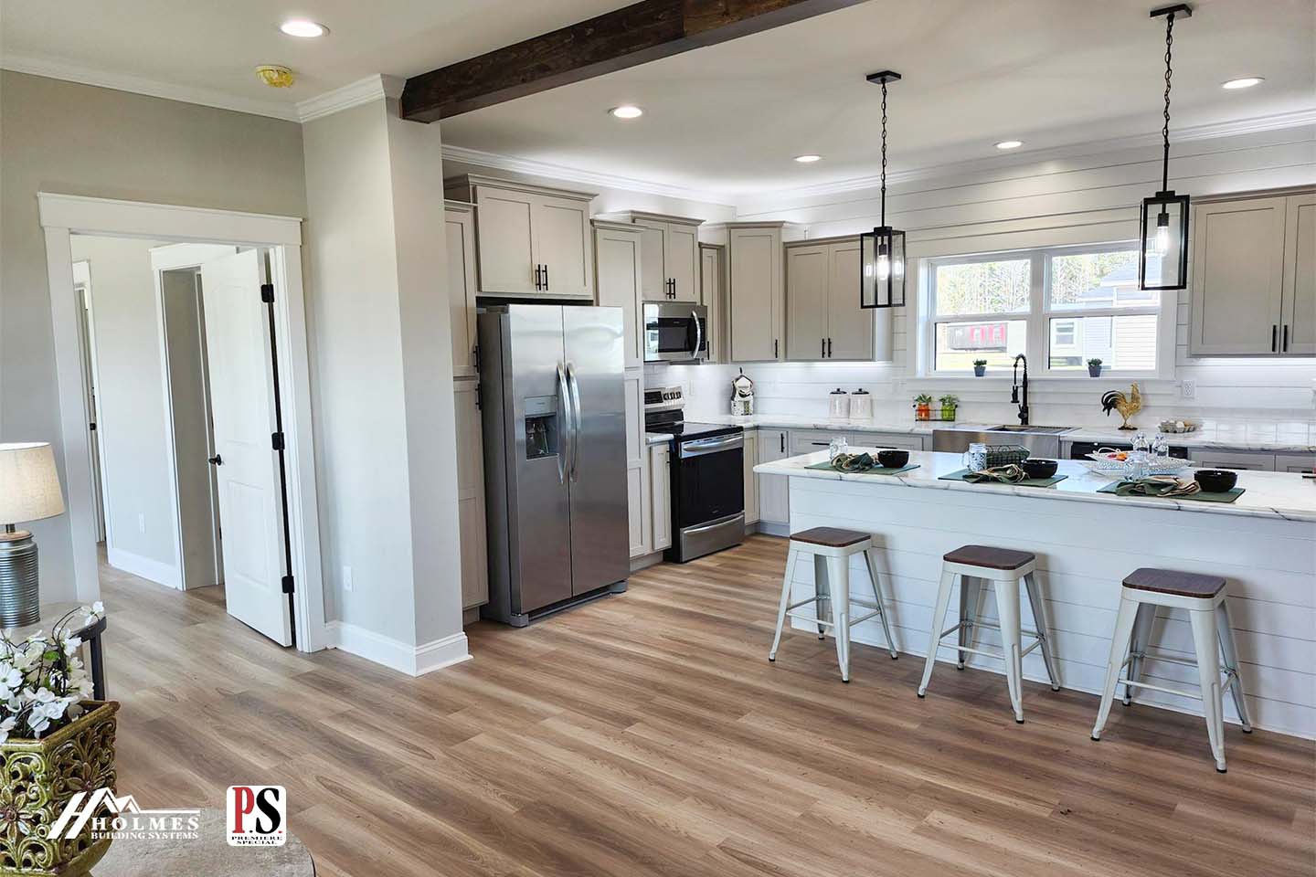 Modern kitchen with beige cabinets and stainless steel appliances, including fridge and stove. A white island with stools is in the center, under two pendant lights. Light wood flooring and a cozy, bright atmosphere.