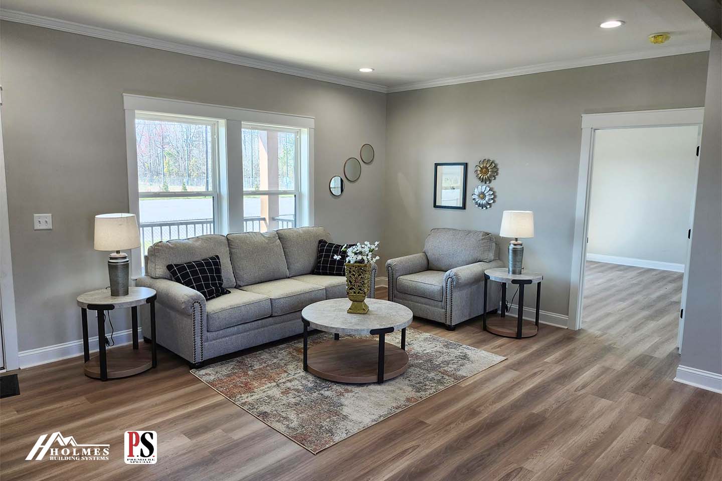 A cozy living room features two gray sofas, a round coffee table on a patterned rug, and three wall mirrors. Natural light streams through large windows.