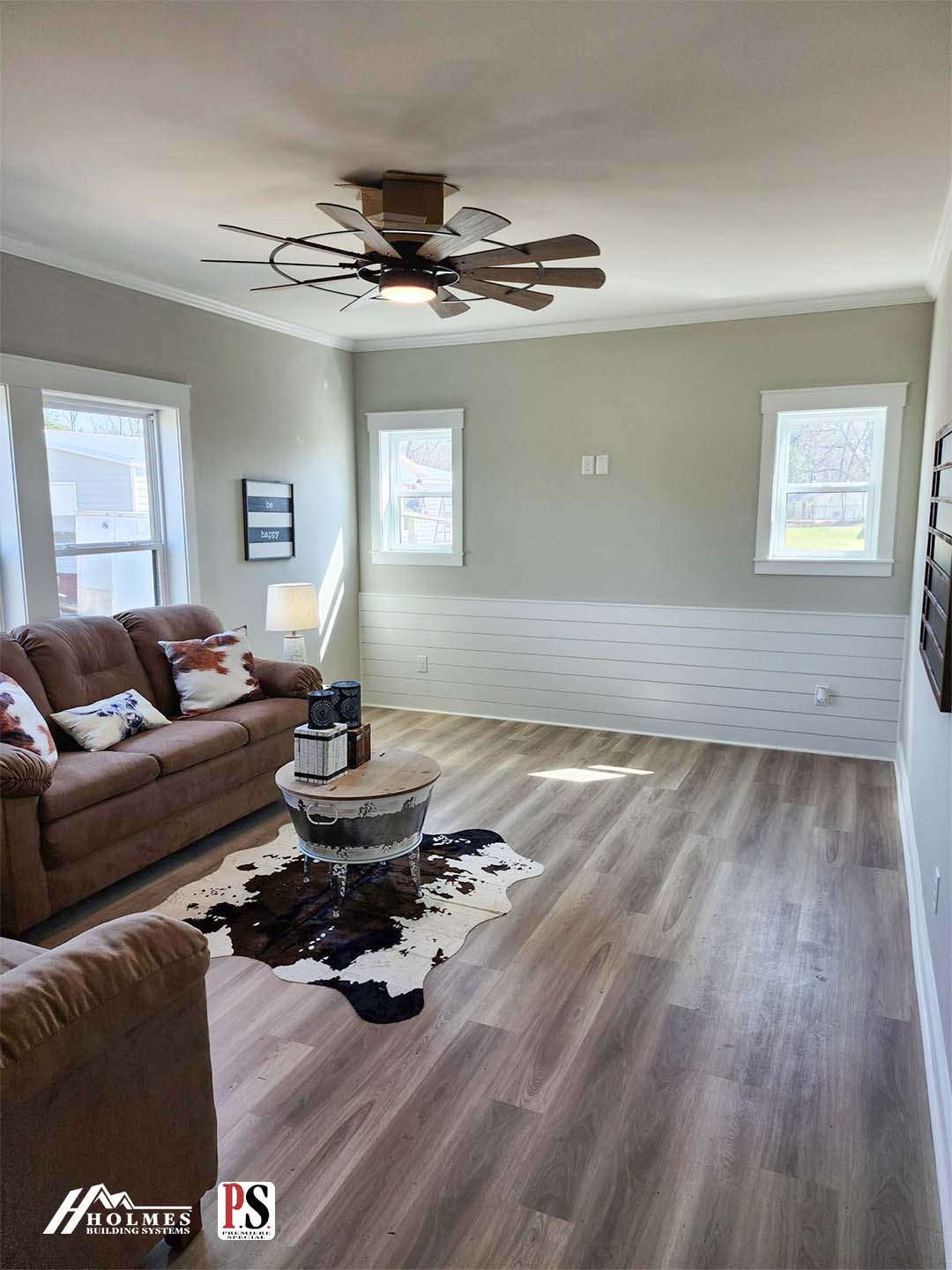 Contemporary living room with brown sofas, neutral walls, and a cowhide rug. Features include large windows, wood flooring, and a ceiling fan. Cozy ambiance.