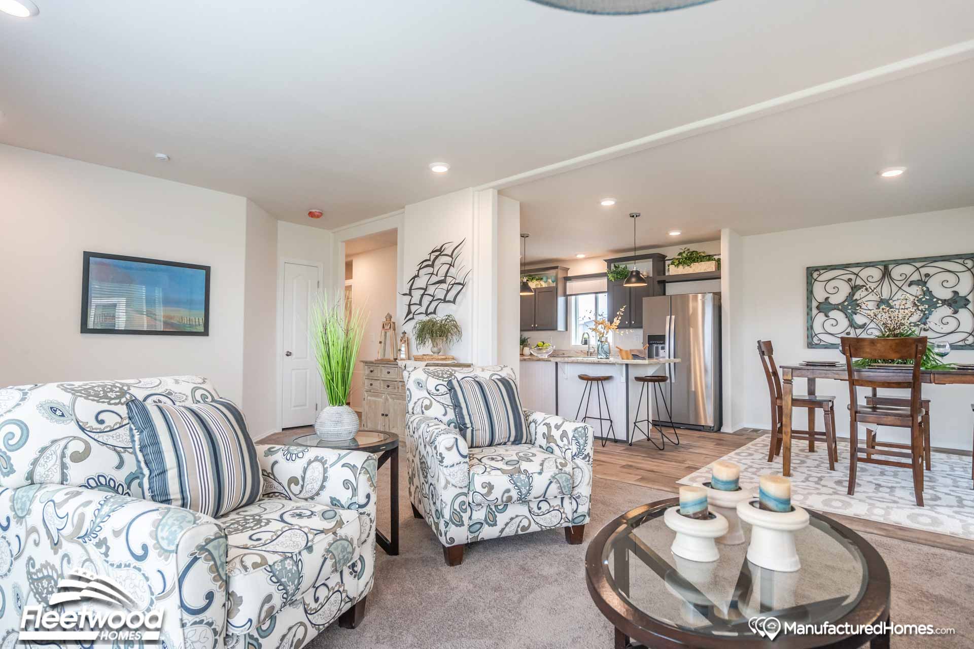 Modern living room with two patterned armchairs, a glass coffee table, and a view into the kitchen. The space is bright and inviting with a cozy, elegant feel.