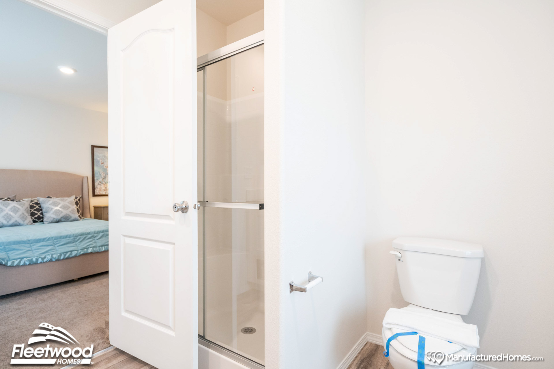 Modern bathroom with glass shower door, white toilet, and neatly folded towels. Bedroom with a made bed and decorative pillows is visible through open door.