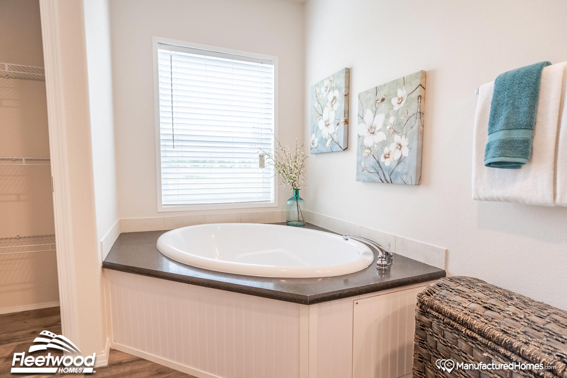 A cozy bathroom corner features an inviting oval bathtub beneath a window with blinds. Floral art decorates the walls, while towels hang nearby for a relaxing ambiance.