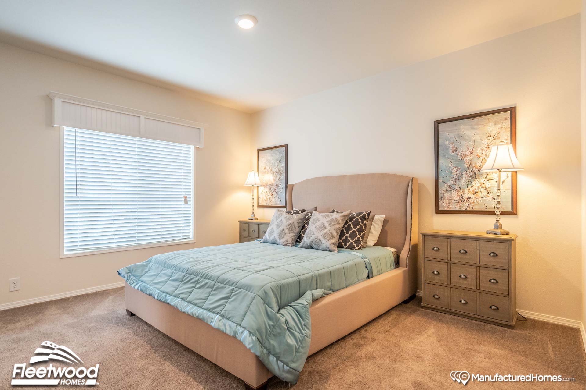 A cozy bedroom with a beige upholstered bed, adorned with pastel bedding and decorative pillows. Two nightstands with lamps and floral art evoke tranquility.