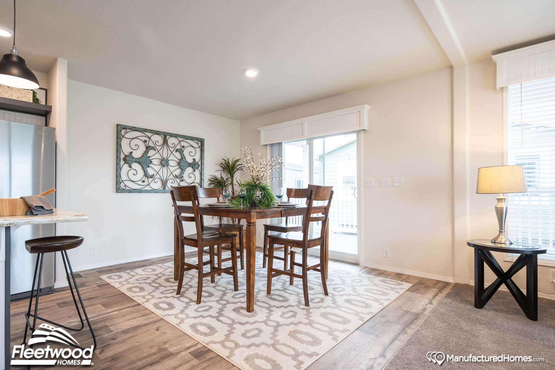 Modern dining room with a wooden table and four chairs on a patterned rug. Bright natural light filters through large windows, creating a cozy ambiance.