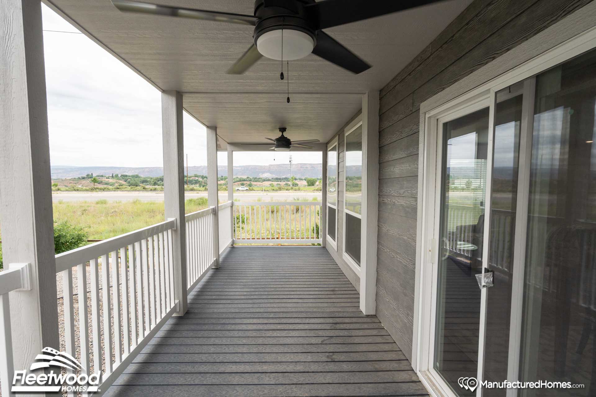 Covered porch with dark wooden floorboards and white railings. Ceiling fans hang from a white roof. Large sliding glass doors open to scenic countryside.