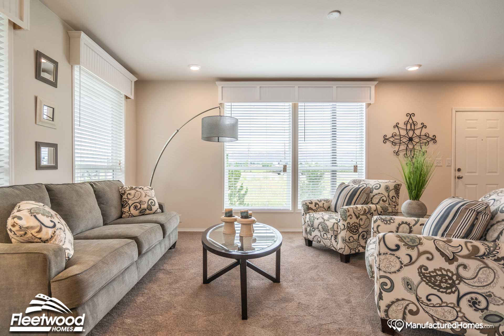 Cozy living room with a gray sofa, patterned armchairs, and a glass coffee table. Large windows brighten the space with natural light, creating a warm atmosphere.