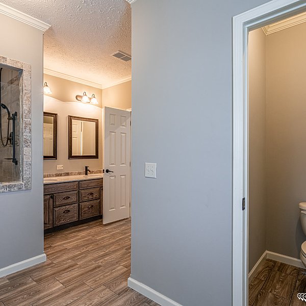 Modern bathroom interior with light gray walls and wood-like flooring. Double vanity with mirrors and warm lighting on the left; a small, open door leads to a toilet on the right. Cozy and elegant ambiance.