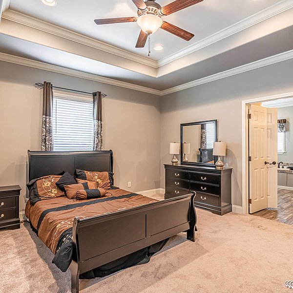 Spacious bedroom with a dark wood sleigh bed, brown and black bedding, and matching furniture. Ceiling fan, soft lighting, and a doorway to a bathroom. Calm and inviting atmosphere.