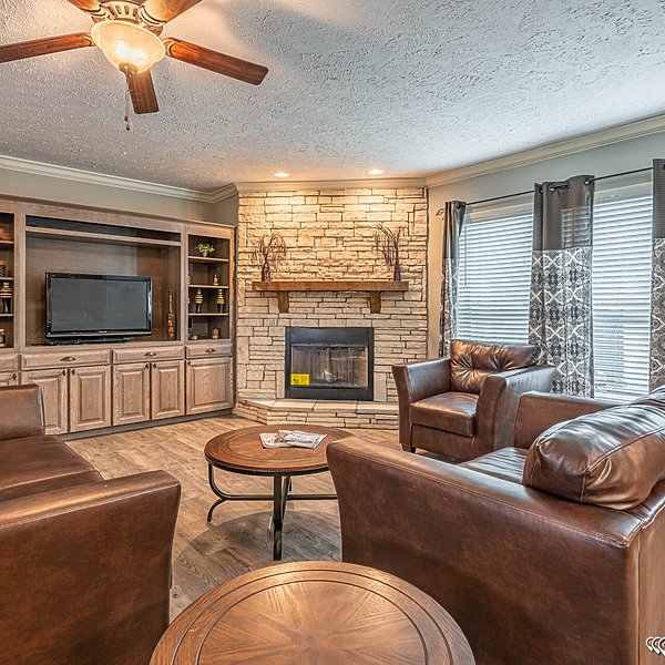 Cozy living room with brown leather chairs and a couch, a wooden coffee table, stone fireplace, built-in TV unit, and warm lighting from a ceiling fan.