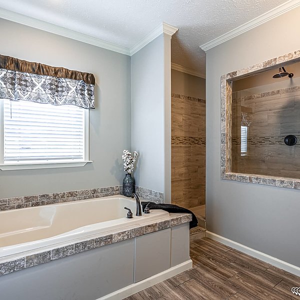 A serene bathroom featuring a white tub with gray stone trim, a dark faucet, and a decorative vase. Beside it, a walk-in shower with beige tiles. The room has light gray walls and wooden flooring with a patterned curtain above the window, conveying a modern, tranquil vibe.