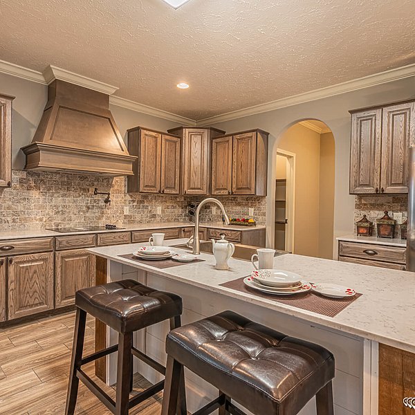Warm-toned kitchen with wooden cabinets and a stone backsplash. Central island features a marble countertop, set with teacups and plates. Two cushioned stools provide seating, creating a cozy, inviting atmosphere.