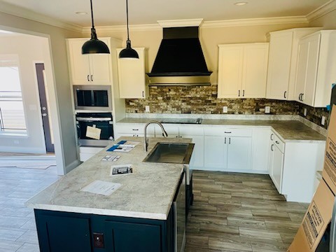 Modern kitchen with white cabinets, black range hood, and stone backsplash. Central island features a sink. Light wood floors and pendant lights add warmth.