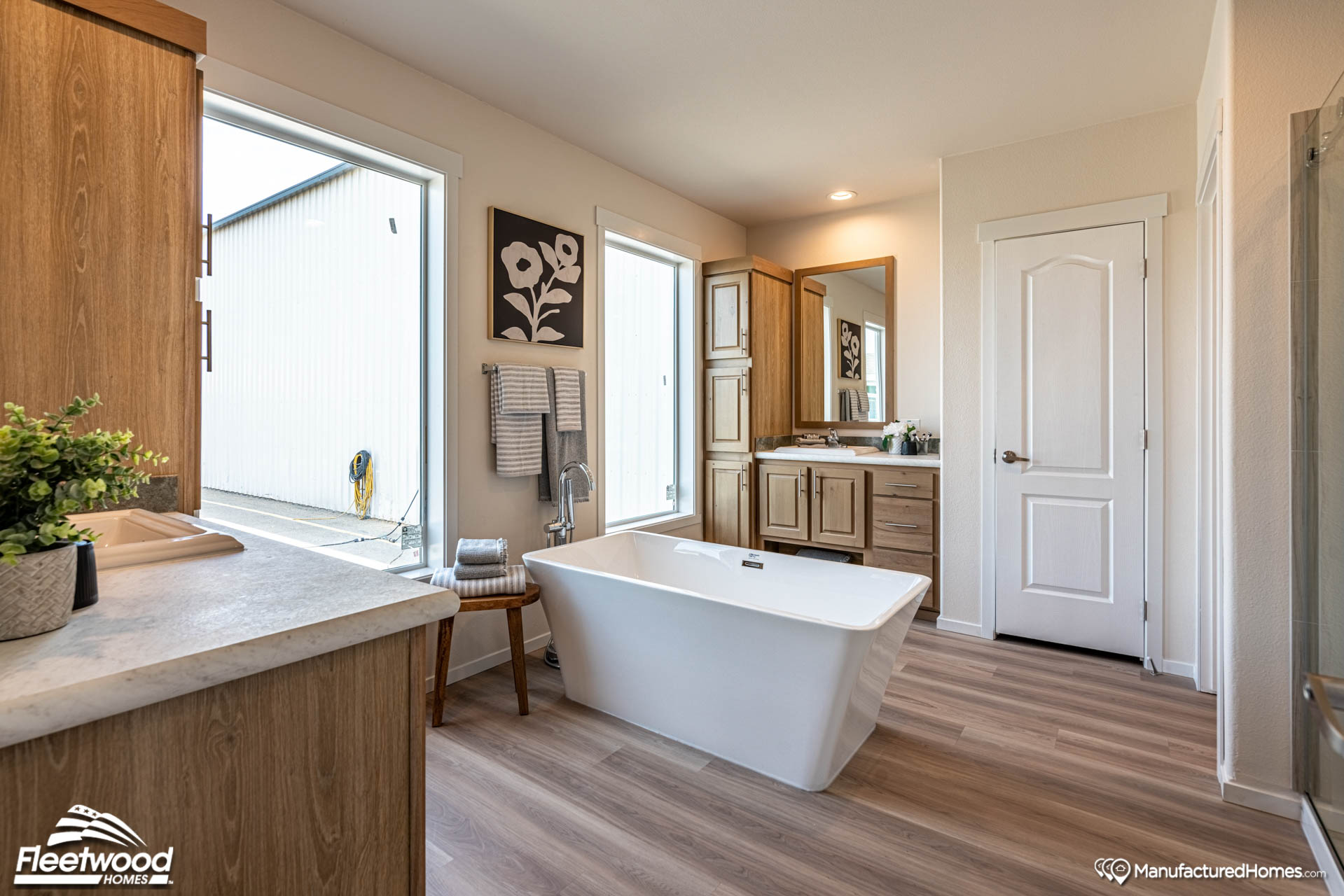 Modern bathroom with light wood cabinetry, freestanding white tub, and large windows. Minimalist decor with plants and artwork, creating a serene atmosphere.