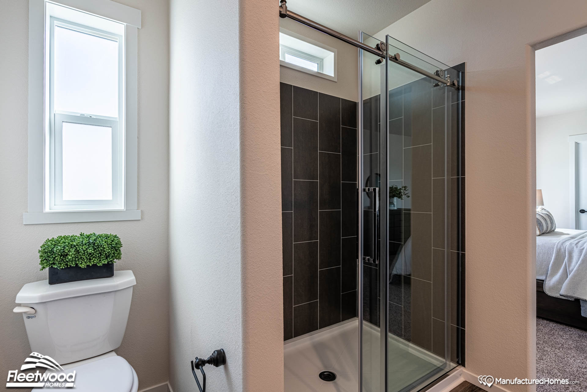 A modern bathroom with a glass shower door and dark tile. There's a window above and a white toilet with a green plant on top, near a cozy bedroom.