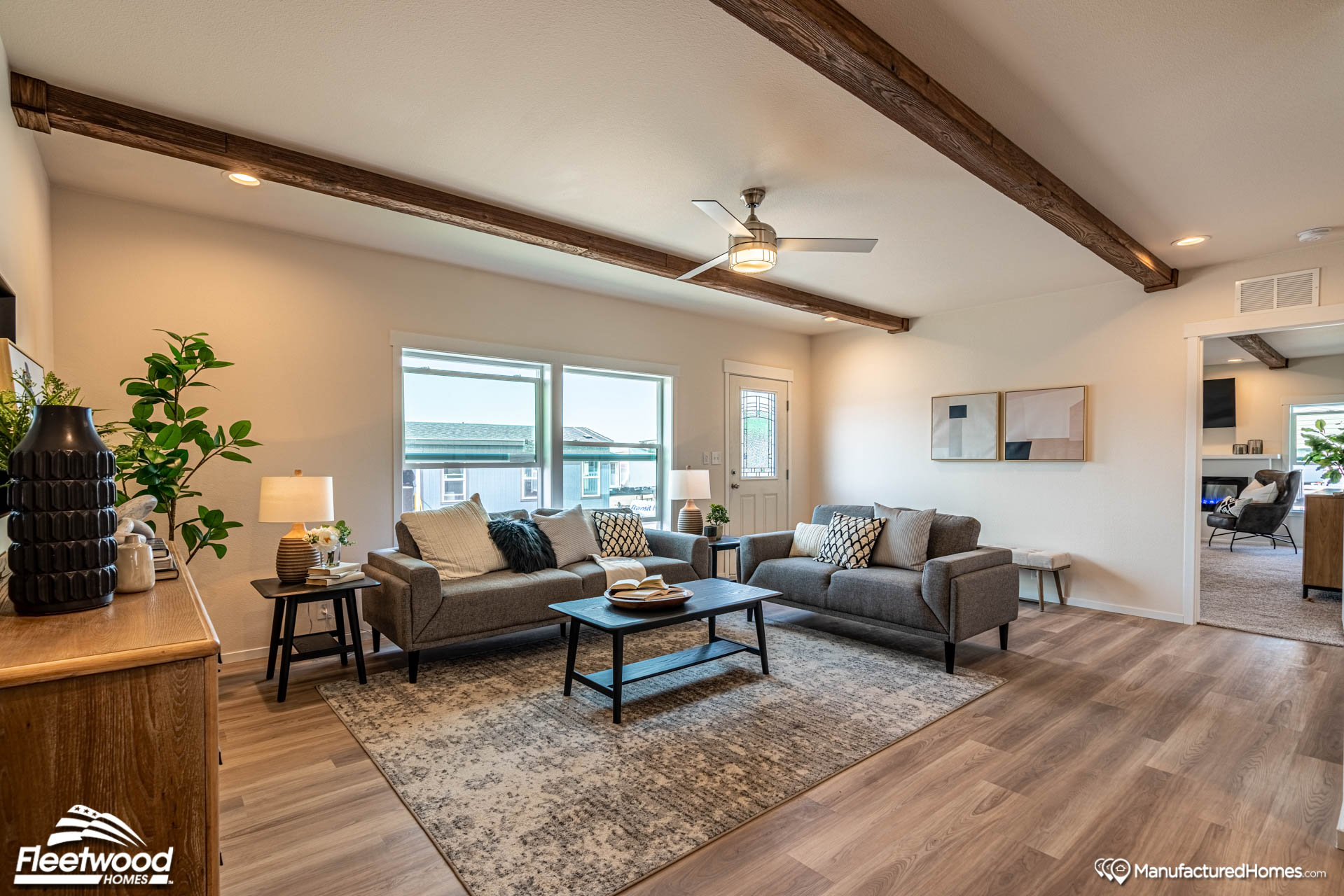 Modern living room with two gray sofas, decorative pillows, a wooden coffee table, and a patterned rug. Large windows and a ceiling fan create a bright, airy feel. Warm lighting and wood beam accents add coziness.