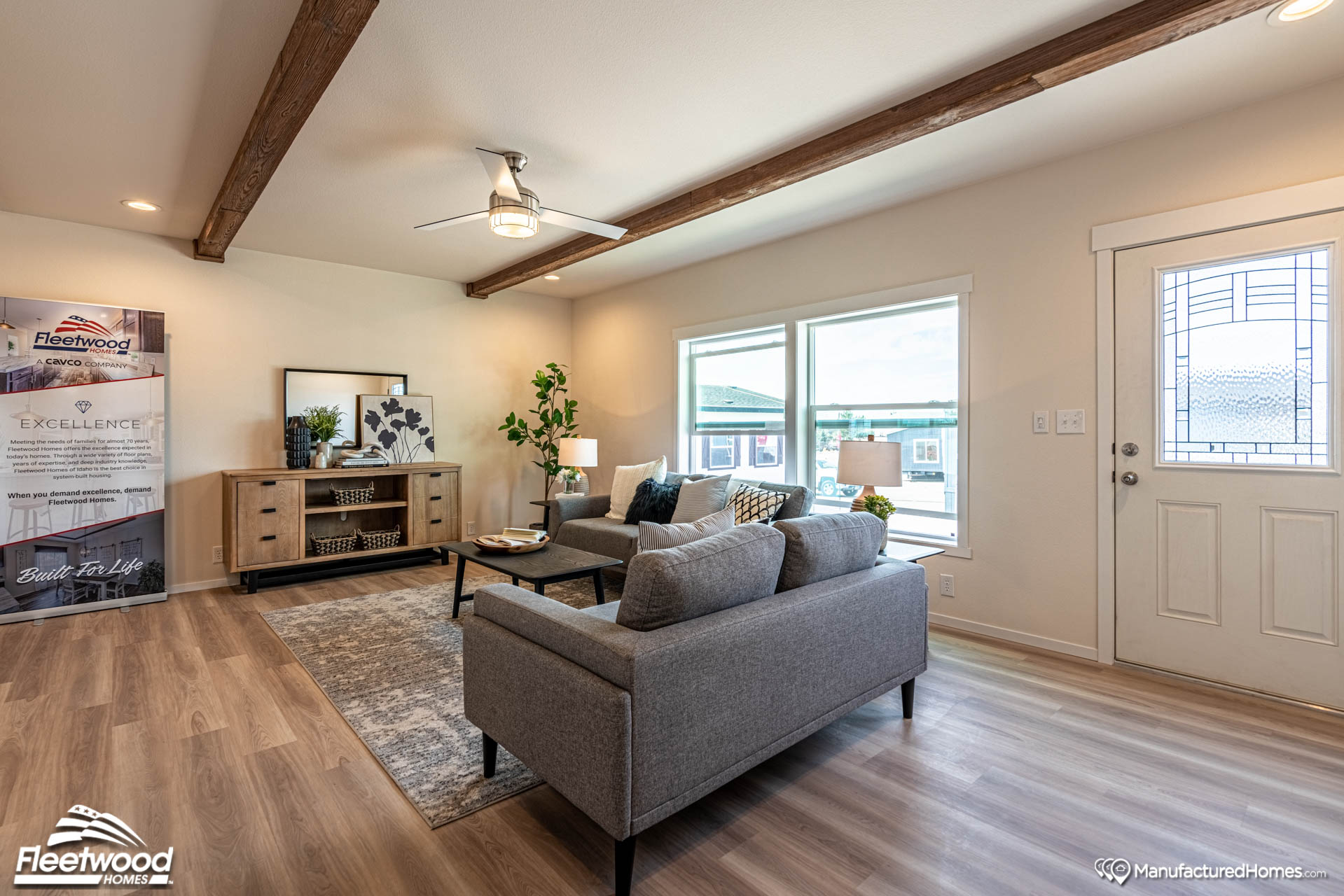 Modern living room with gray sofas, a wooden coffee table, and a media console on wood floors. Natural light streams through large windows, highlighting a cozy, inviting ambiance.