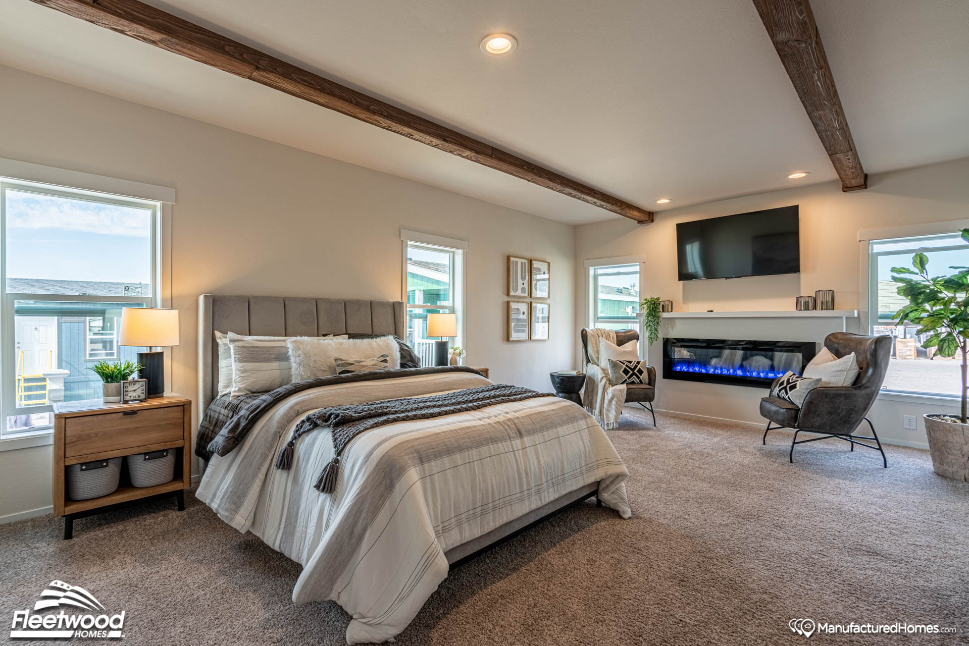 Spacious bedroom with a cozy bed, wooden beams, and large windows. Features a modern fireplace, wall-mounted TV, plush chairs, and soft, neutral tones.