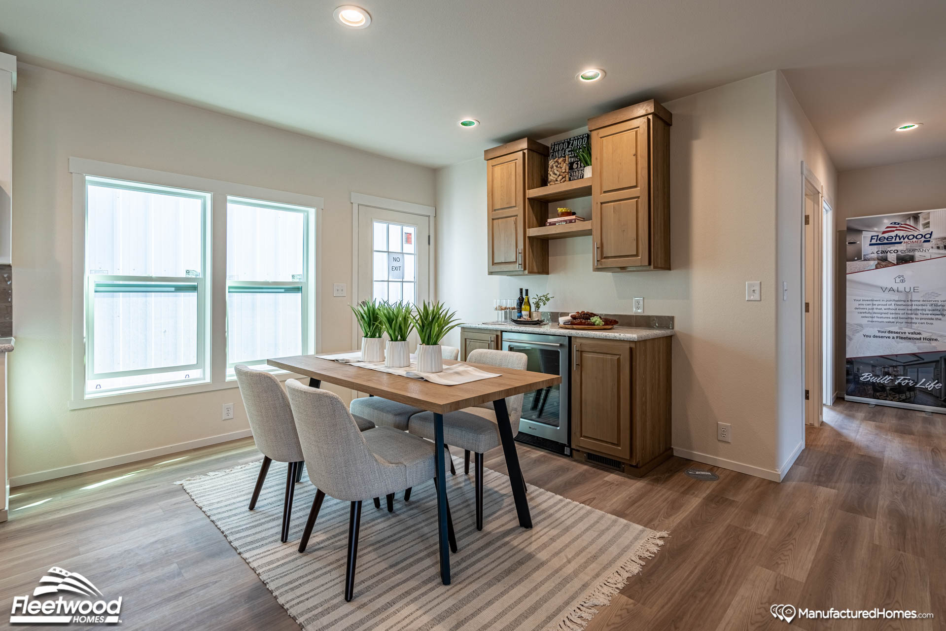 Modern dining area with a wooden table and four chairs on a striped rug. Wooden cabinets and decor create a cozy, inviting atmosphere. Bright natural lighting.