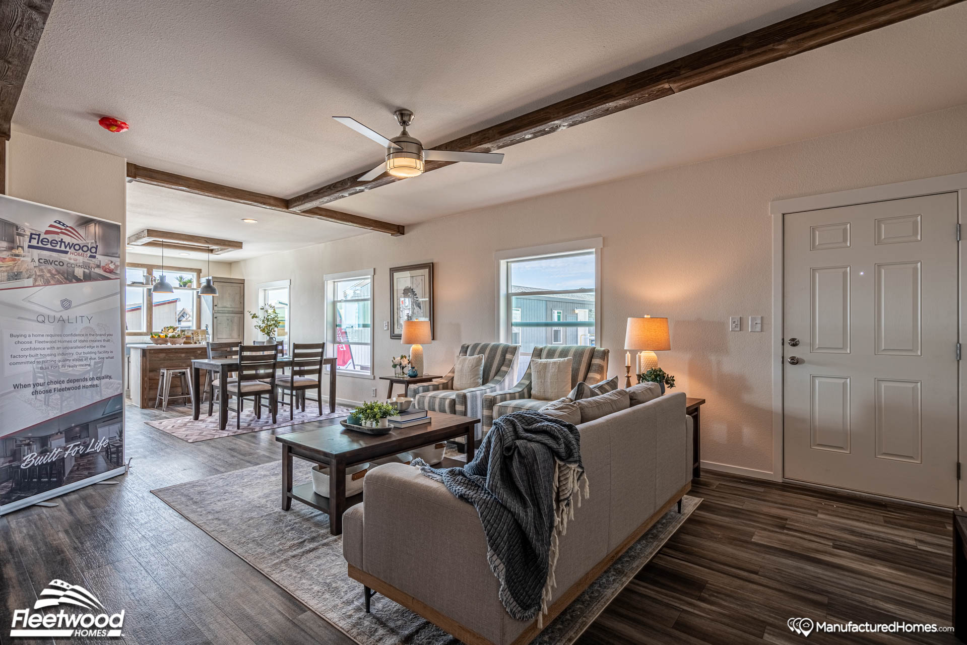 Modern open-concept living room with neutral tones, featuring a cozy seating area, wooden beams, large windows, dining table, and warm lighting.