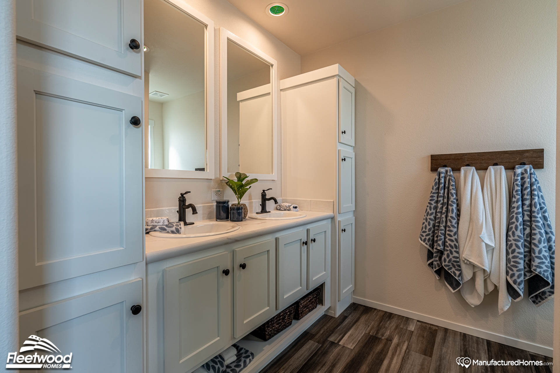 A modern bathroom featuring a white double sink vanity with black faucets, two mirrors, and a tall cabinet. Towels hang on a wall rack; wood flooring adds warmth.