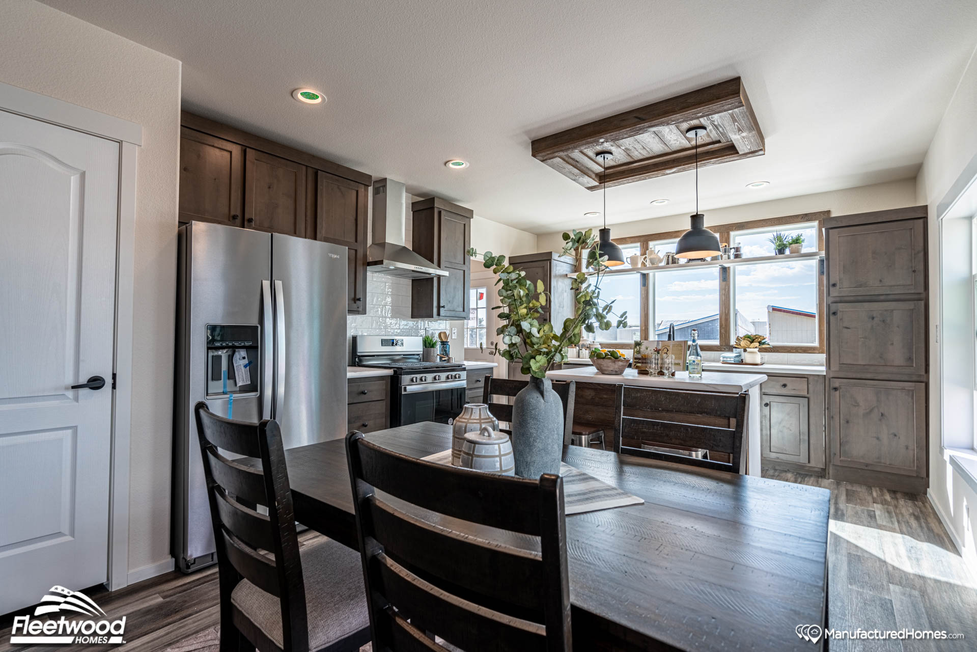 Modern kitchen with dark wood cabinets and a stainless steel fridge. A dining table with a vase of flowers is in the foreground, and large windows let in natural light.