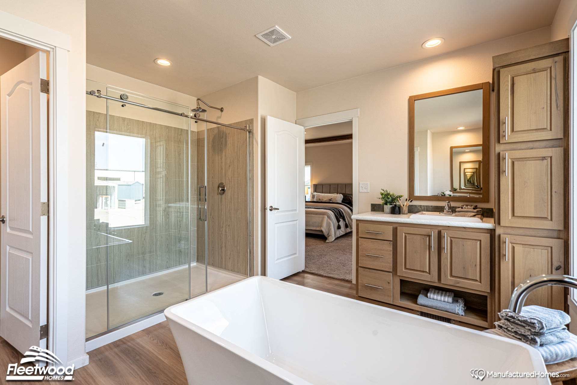 Modern bathroom featuring a freestanding tub, glass-enclosed shower, wooden vanity with a large mirror, and view into a cozy bedroom. Natural tones create a relaxing atmosphere.