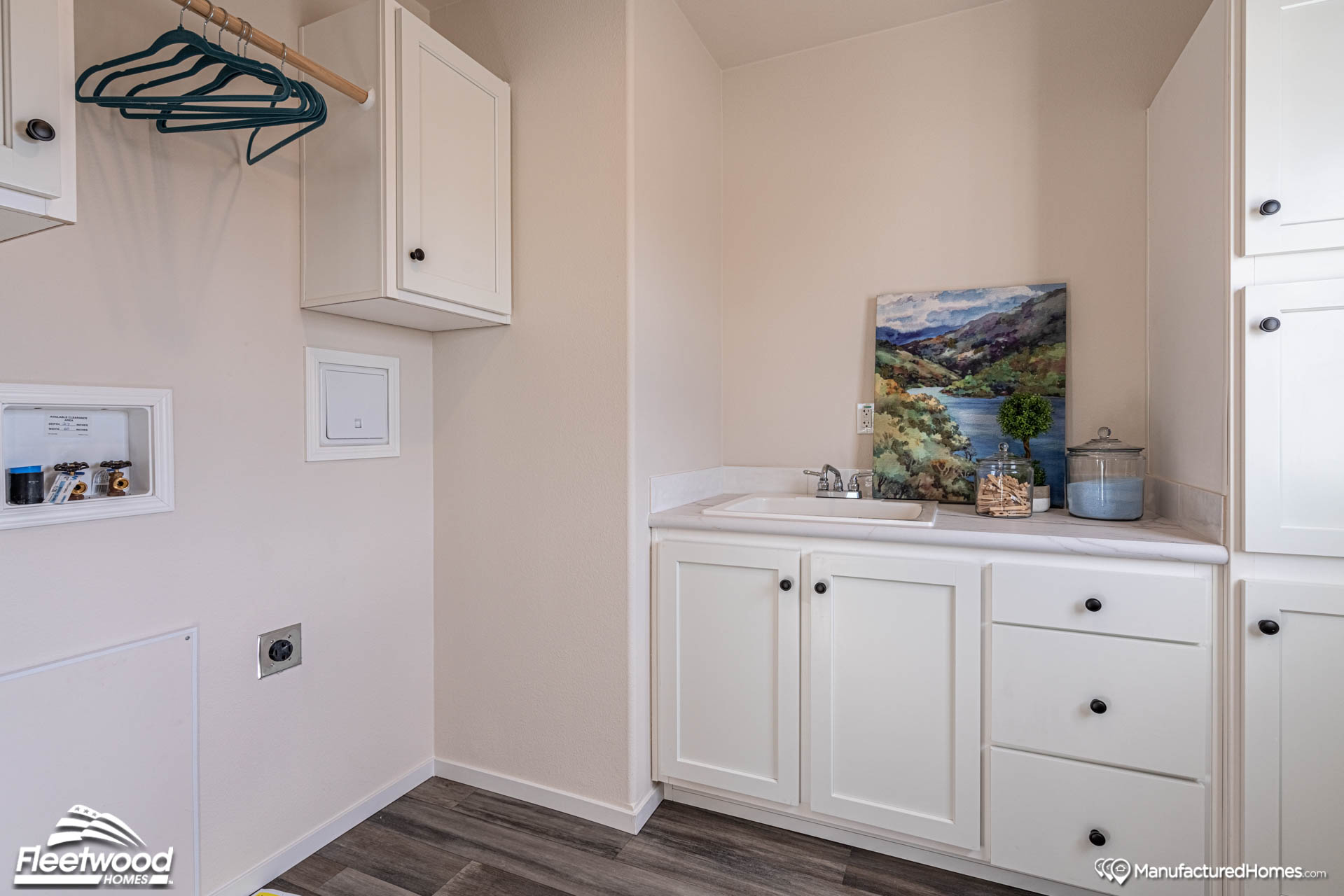 Laundry room with white cabinets, a small sink, and a countertop. Blue and green hangers on a rod, landscape painting, and storage items add color.