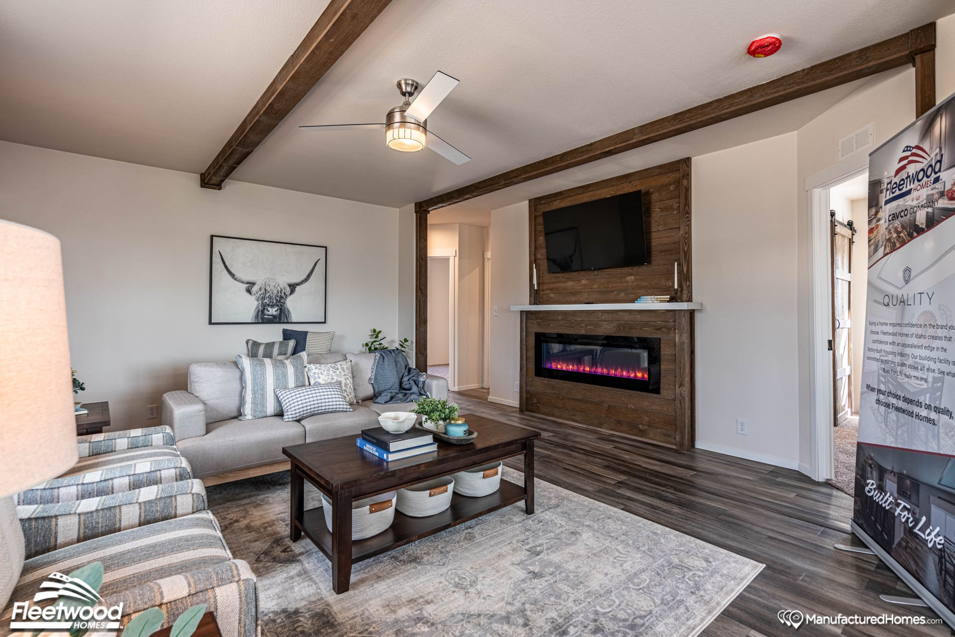 Modern living room with wood beams, cozy sofa, armchair, and a cow artwork. Features a fireplace, TV, and a coffee table with books. Warm, inviting atmosphere.