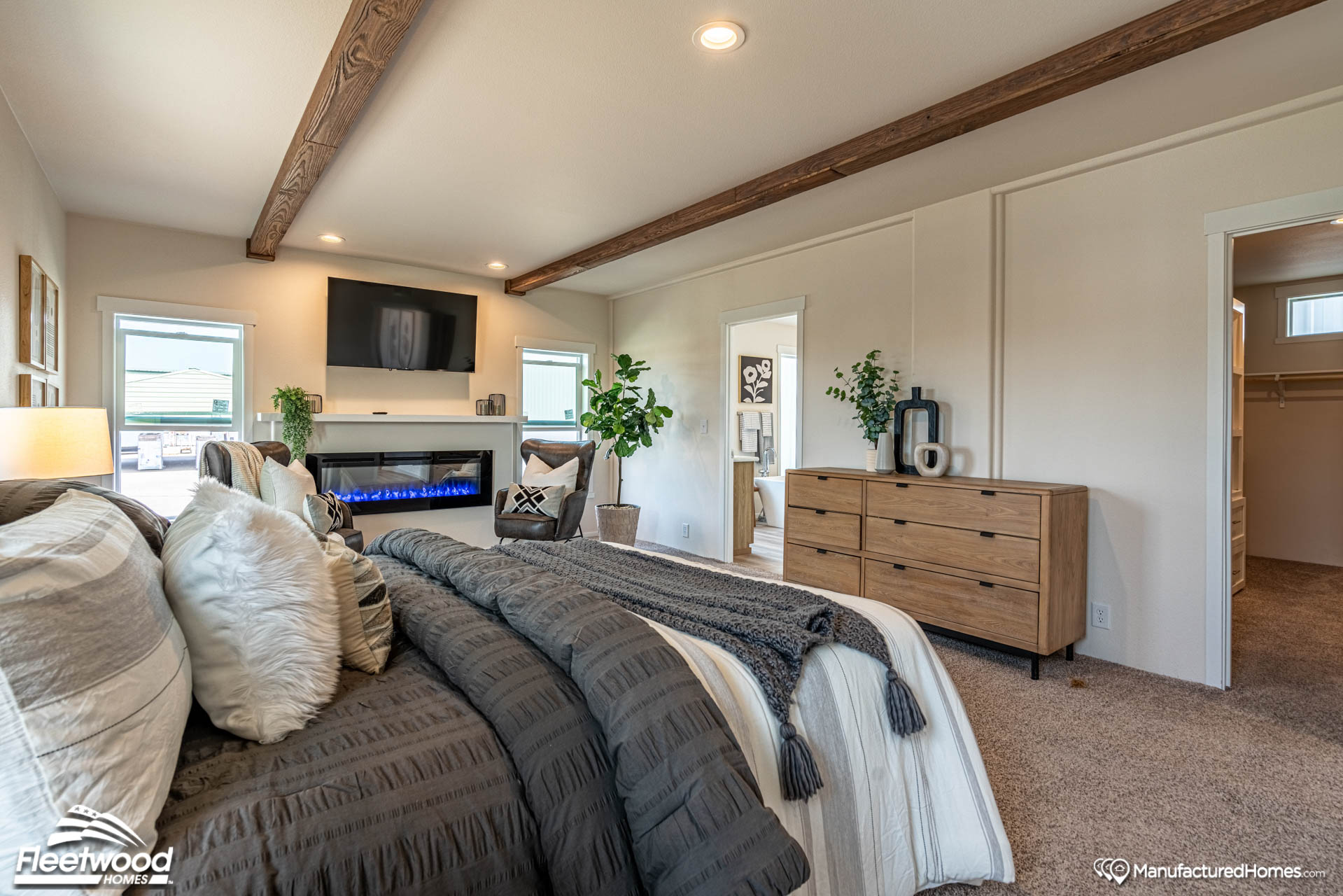 A cozy bedroom features a plush, neatly made bed with patterned pillows. A modern fireplace and TV are set on the opposite wall. Wooden beams and plants add warmth.