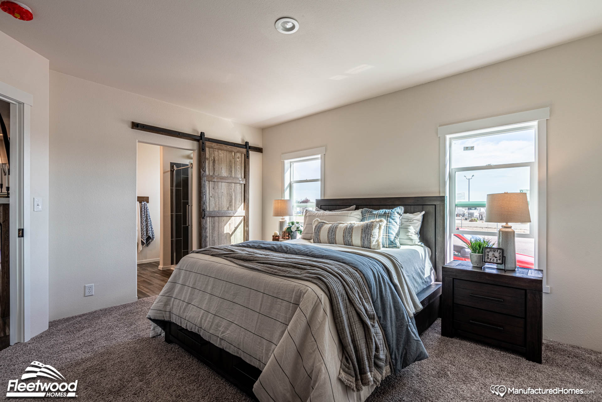 A cozy bedroom with a neatly made bed features gray and blue bedding. Two windows flank the headboard, inviting natural light. A rustic sliding barn door opens into an adjacent room. A warm, inviting atmosphere is enhanced by bedside lamps on dark wood nightstands.