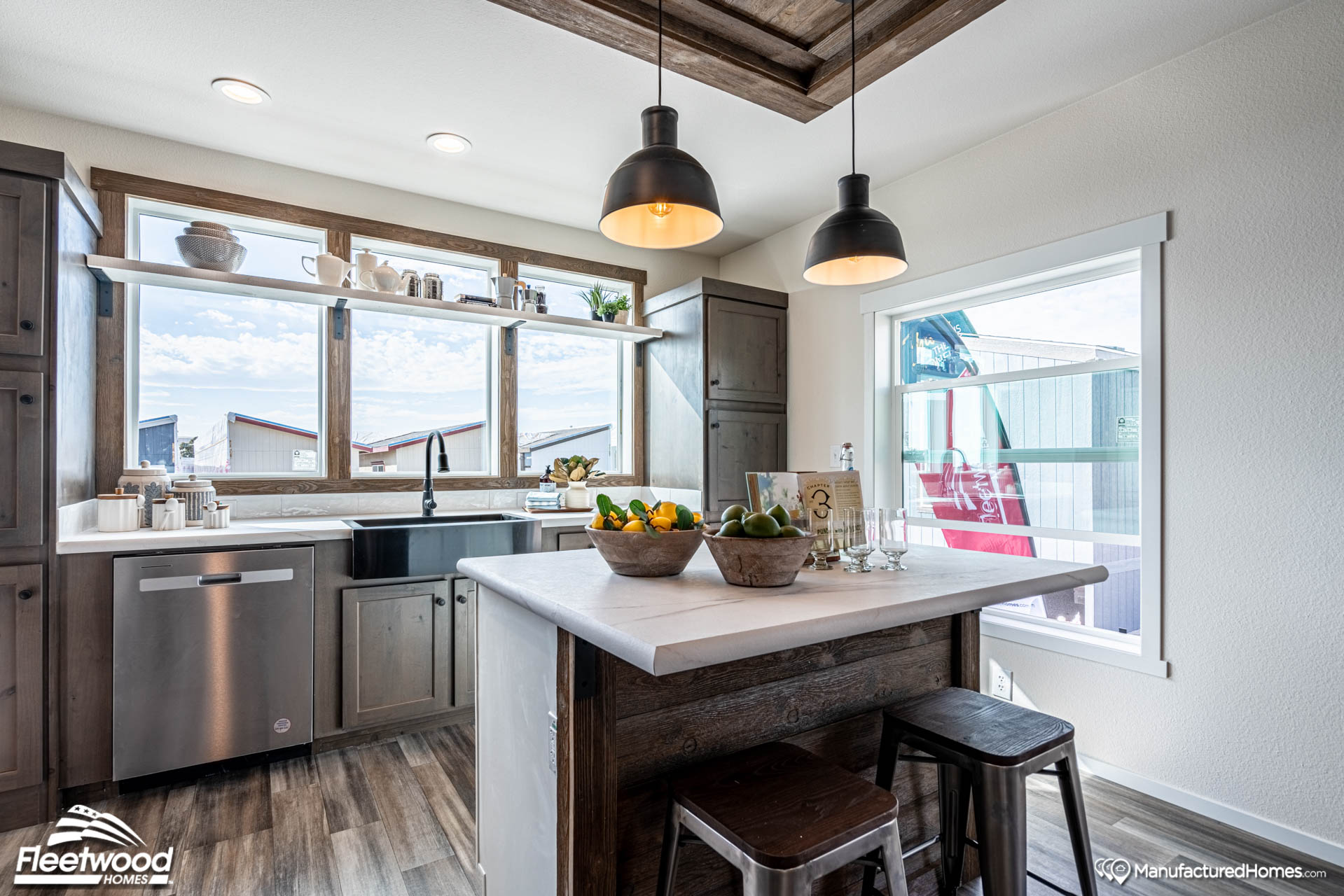 Modern kitchen with white walls and wood floor, featuring an island with fruit bowls and wooden stools. Large windows offer bright, natural light.