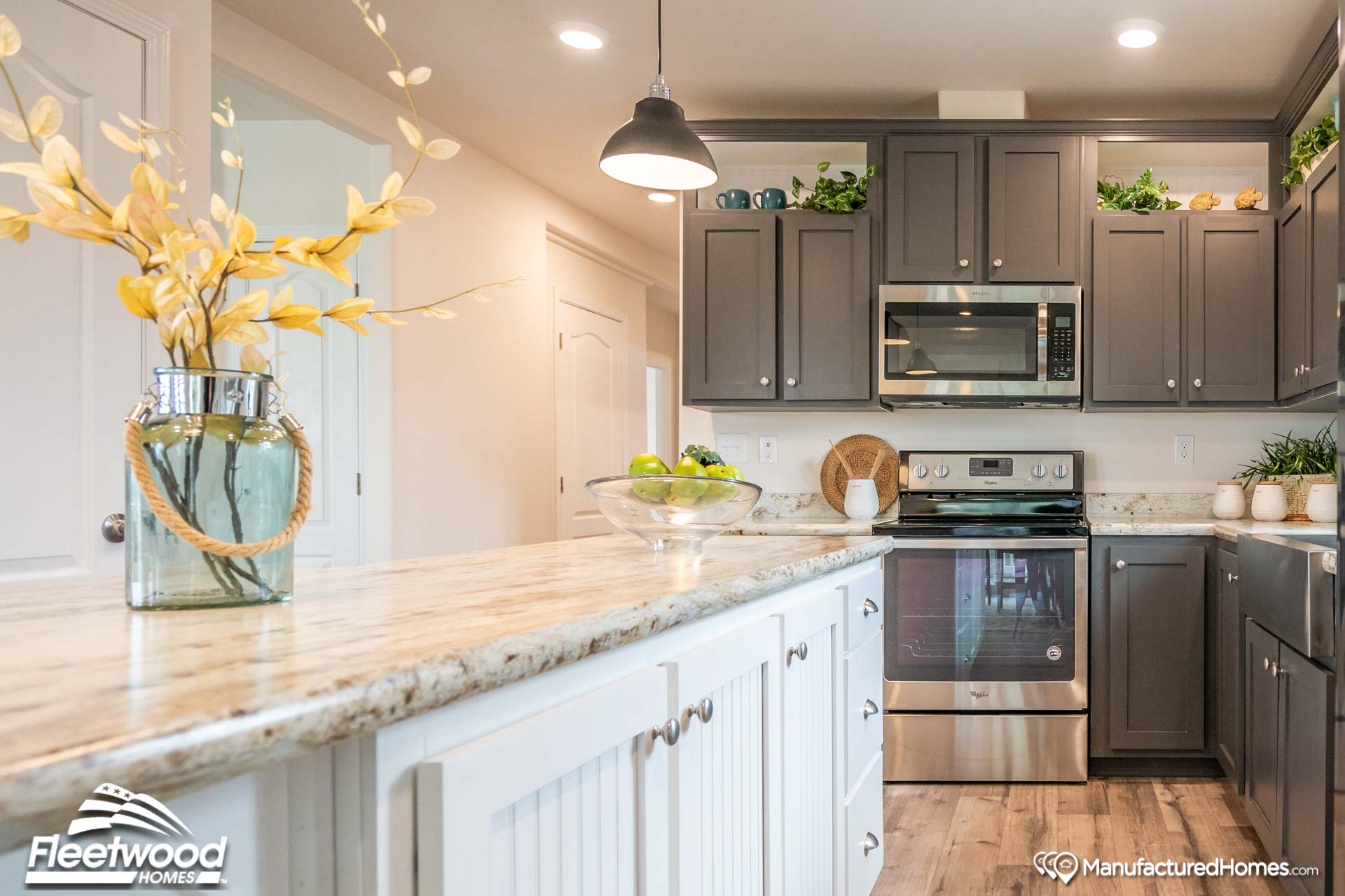 Bright kitchen with gray cabinets, a stainless steel stove, and white countertops. Decor includes a jar of yellow leaves and a bowl of green apples.