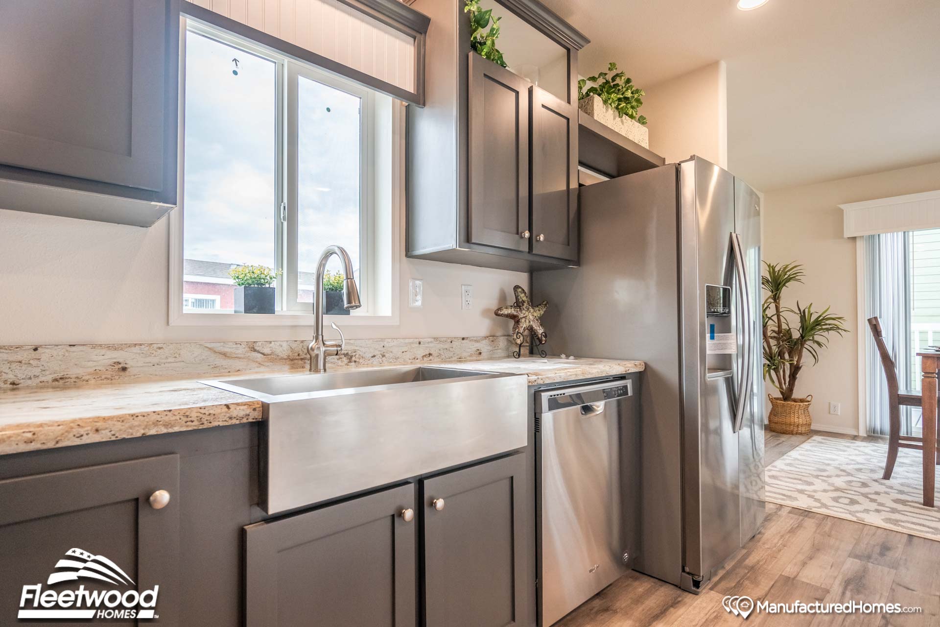 Modern kitchen with gray cabinets, stainless steel appliances, and a farmhouse sink under a window. Decor includes potted plants, creating a fresh, clean atmosphere.
