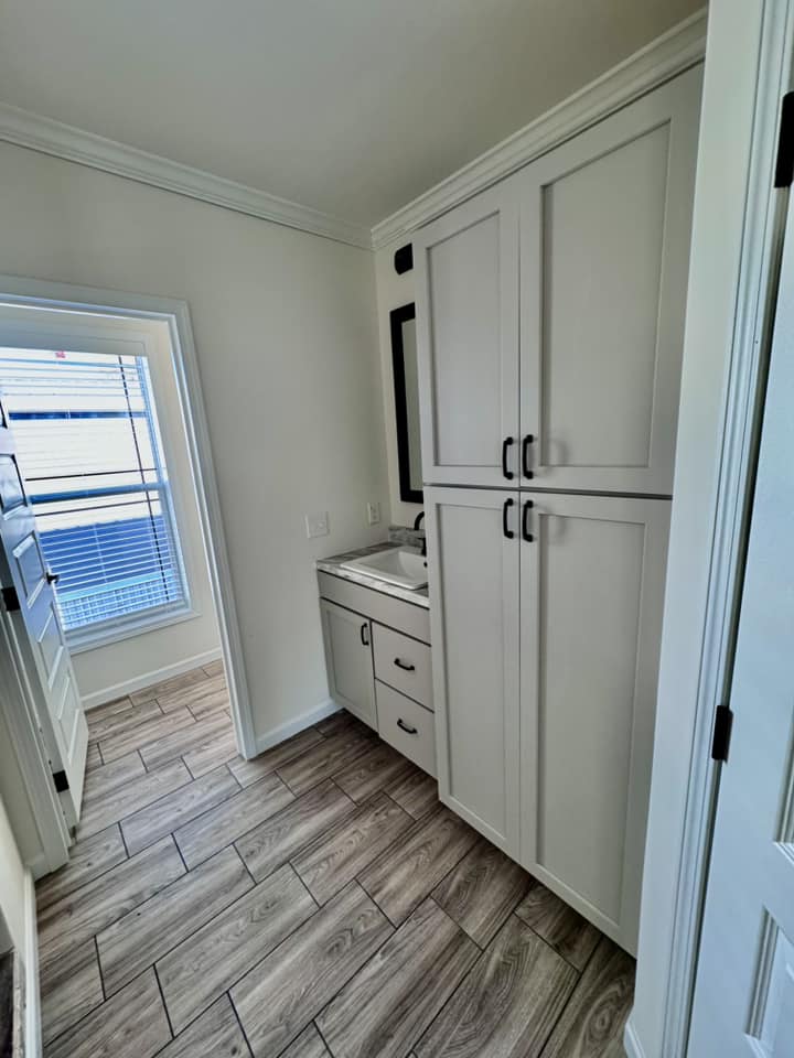 Narrow laundry room with light wooden floors, white cabinets, and a small sink. A large window on the left lets in natural light, creating a bright atmosphere.